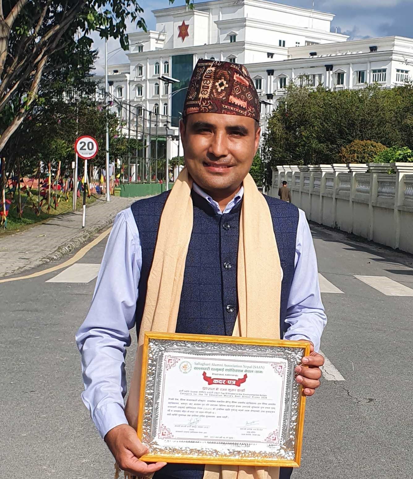 A man wearing a traditional Nepali cap and scarf stands on a street, smiling and holding a framed certificate. A large white building and a speed limit sign are visible in the background.