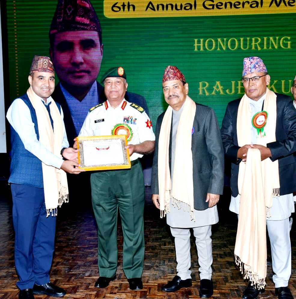 Four men in traditional Nepali attire and scarves stand on a stage, with one military officer giving a framed certificate to another man. A large screen with text serves as the backdrop.