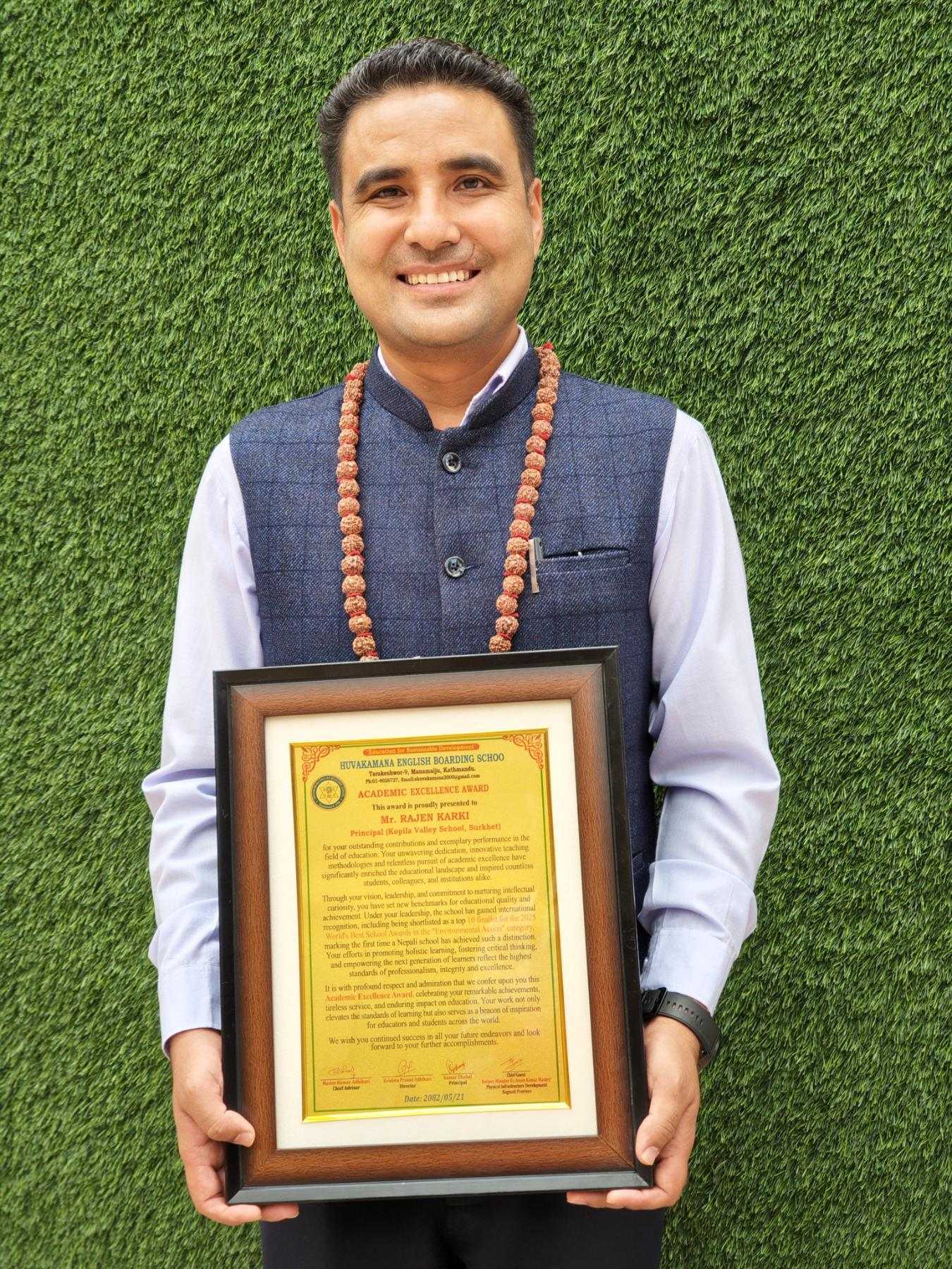 A smiling man in a blue vest and white shirt, wearing a bead garland, stands in front of a green textured background holding a framed certificate or plaque.