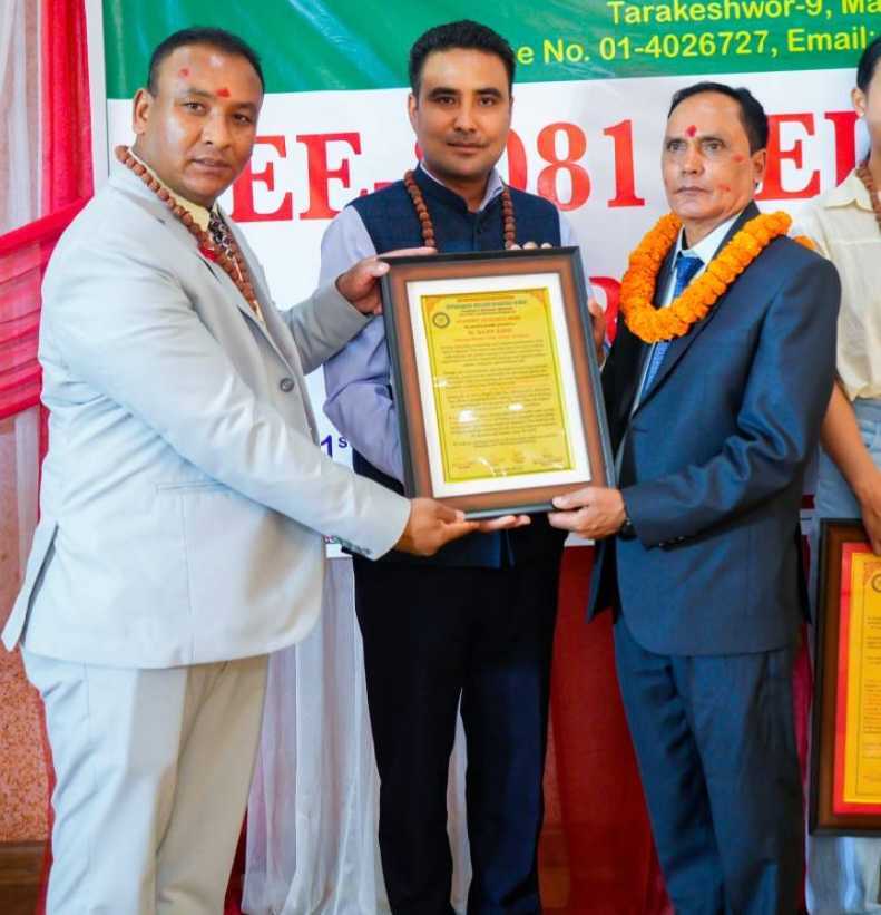 Three men in formal attire, wearing orange flower garlands and red forehead marks, pose together. Two of them hold a framed certificate while standing in front of a decorative backdrop with text.