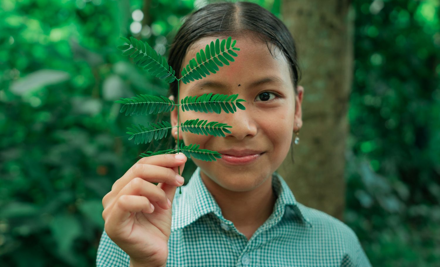 A young girl in a green checkered shirt stands outdoors, smiling and holding a leafy fern branch in front of one eye. The lush, green foliage of the forest forms a blurred background.