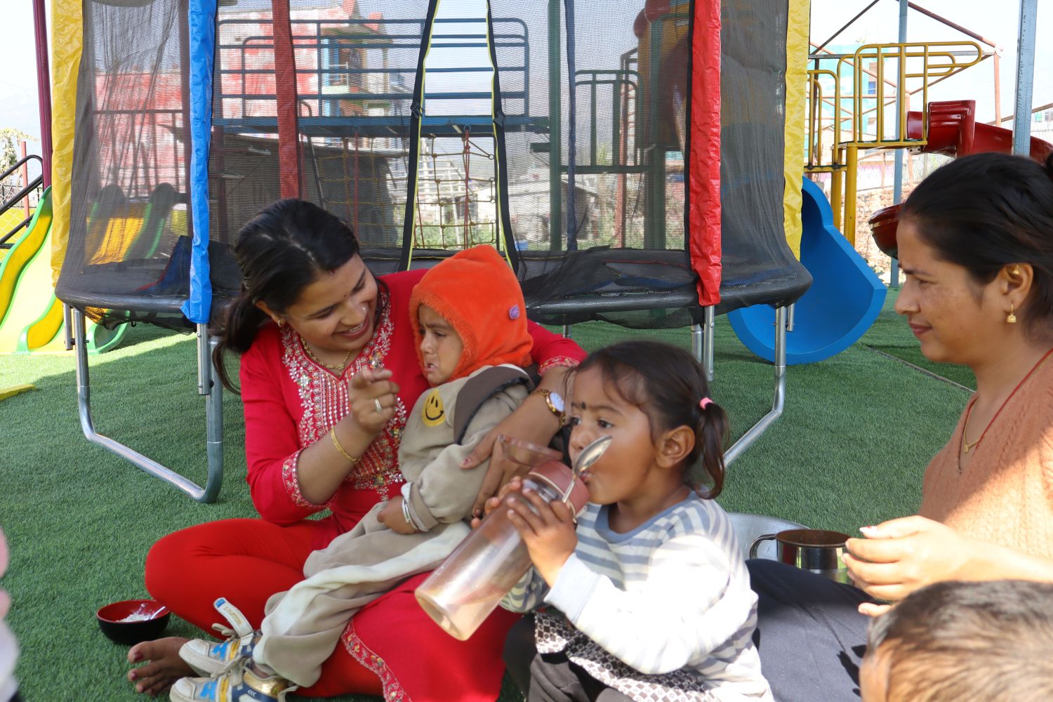 Two women and two children sit on artificial grass at a playground. One woman helps a child in an orange hat, while the other child drinks from a metal cup. Playground equipment is visible in the background.