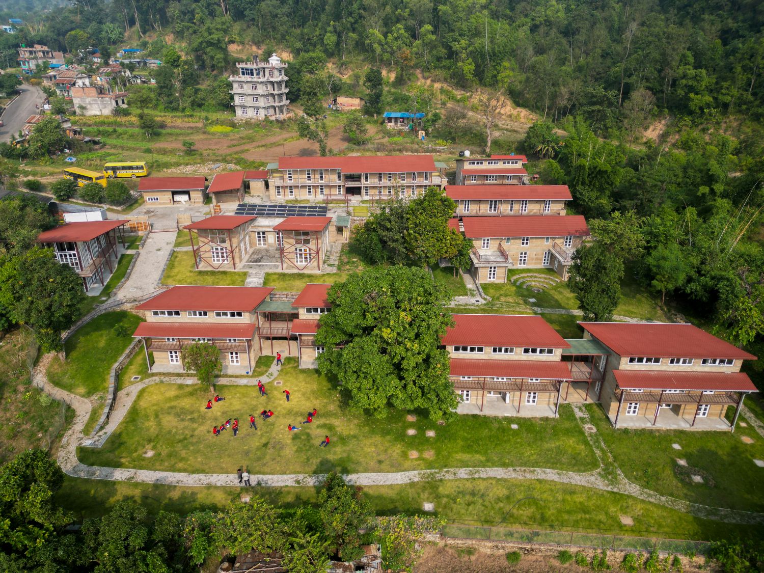 Aerial view of a school complex with red-roofed buildings surrounded by greenery; students in red uniforms play on a grassy field, with trees and a hilly forest in the background.