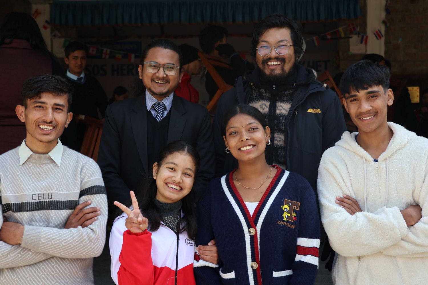 A group of six people, four men and two women, stand close together outdoors, smiling at the camera. One woman flashes a peace sign. They are casually dressed and appear happy and friendly.