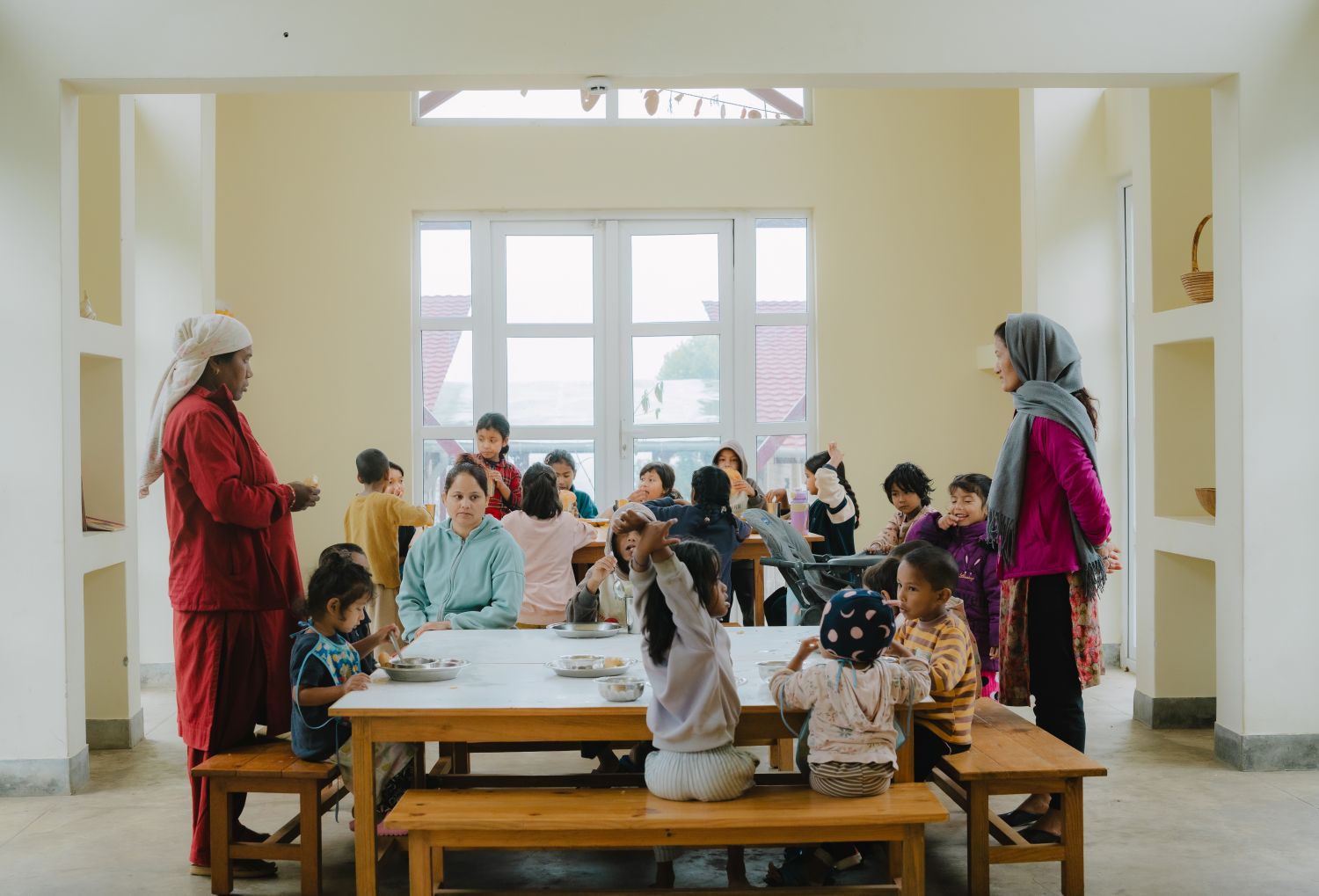 Children sit around wooden tables for a meal, in a room filled with windows and light. Two women stand at each end of the table supervising.