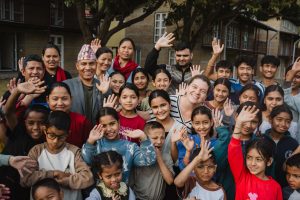 A group of children wave at the camera, with a few adults in the middle.