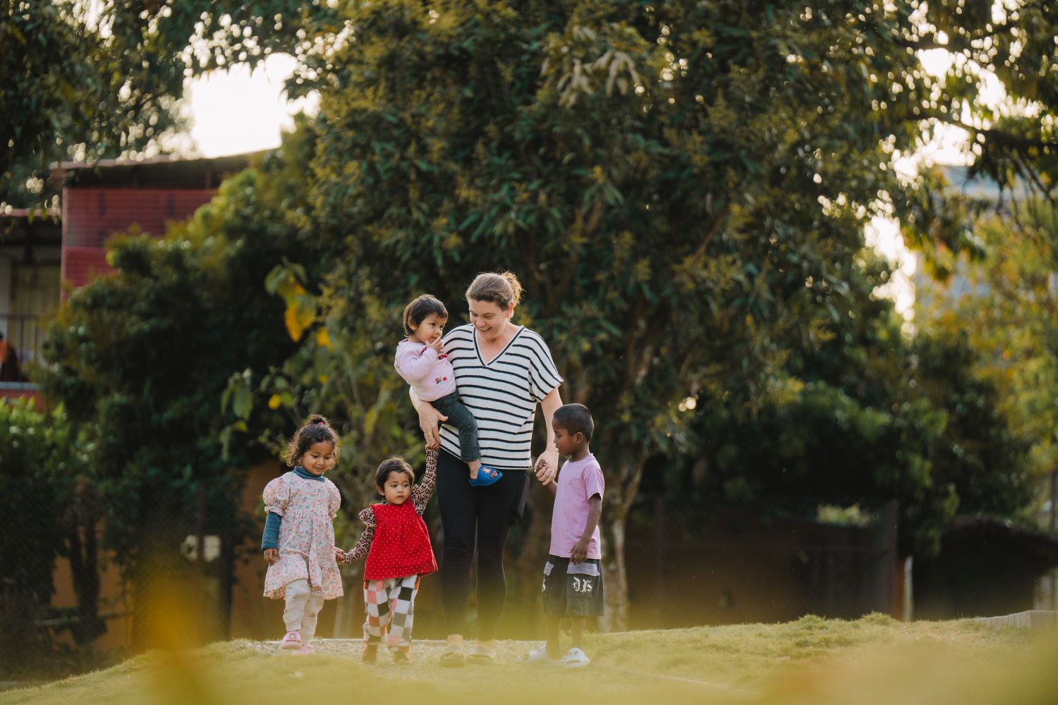 A woman walks with young children in a field with trees behind them.