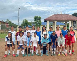 A group of girls in soccer uniforms poses on a dirt field.