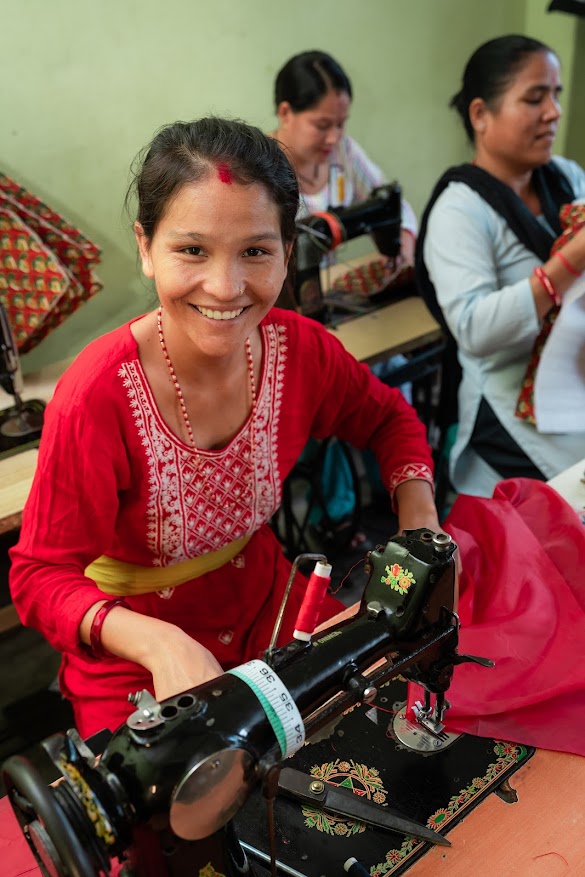 A woman in a red dress sits at a sewing machine and is smiling.