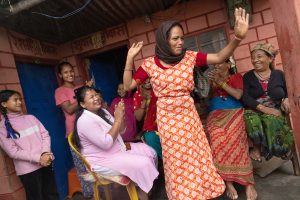 A group of women sit in a half circle while another woman dances in the middle. Some women are clapping, and all of them look happy.