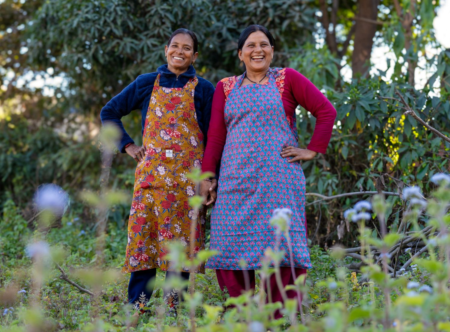 Two women stand in a field wearing handmade aprons, one orange/earthtones and one purple. They are smiling and look happy.