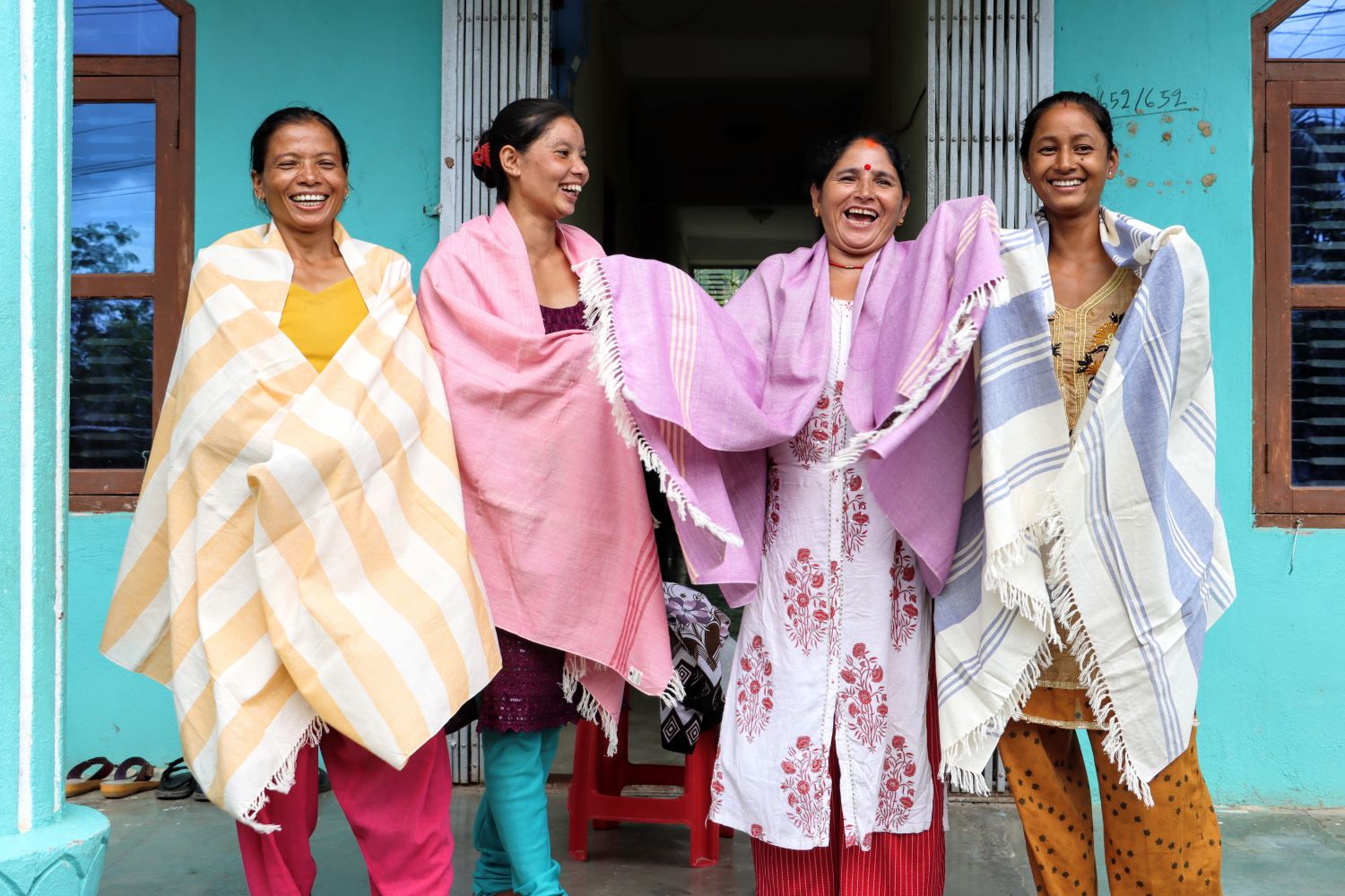 Four women stand in front of a turquoise building, smiling and laughing. They are wearing handmade shawls of various colors.