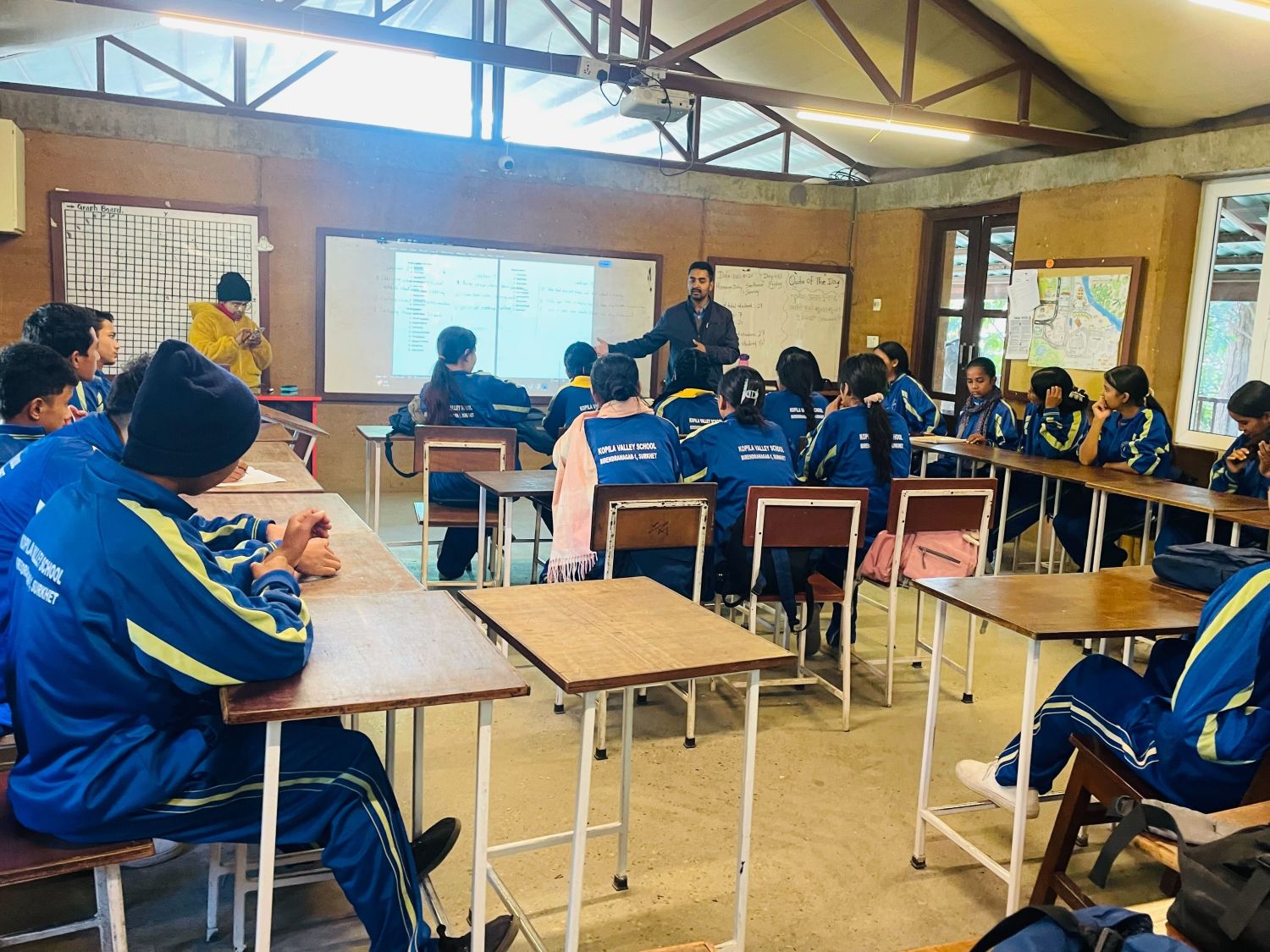 A group of students sit at desks while a teacher stands at the front of the classroom.