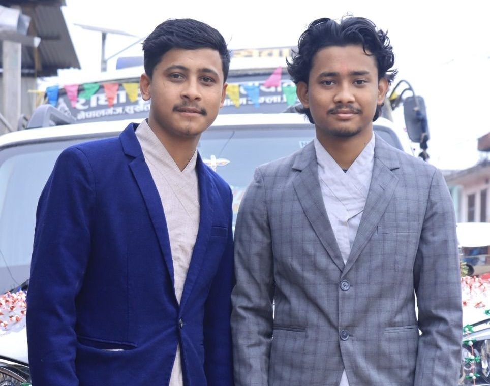 Two young men wearing suit jackets pose on a city street.