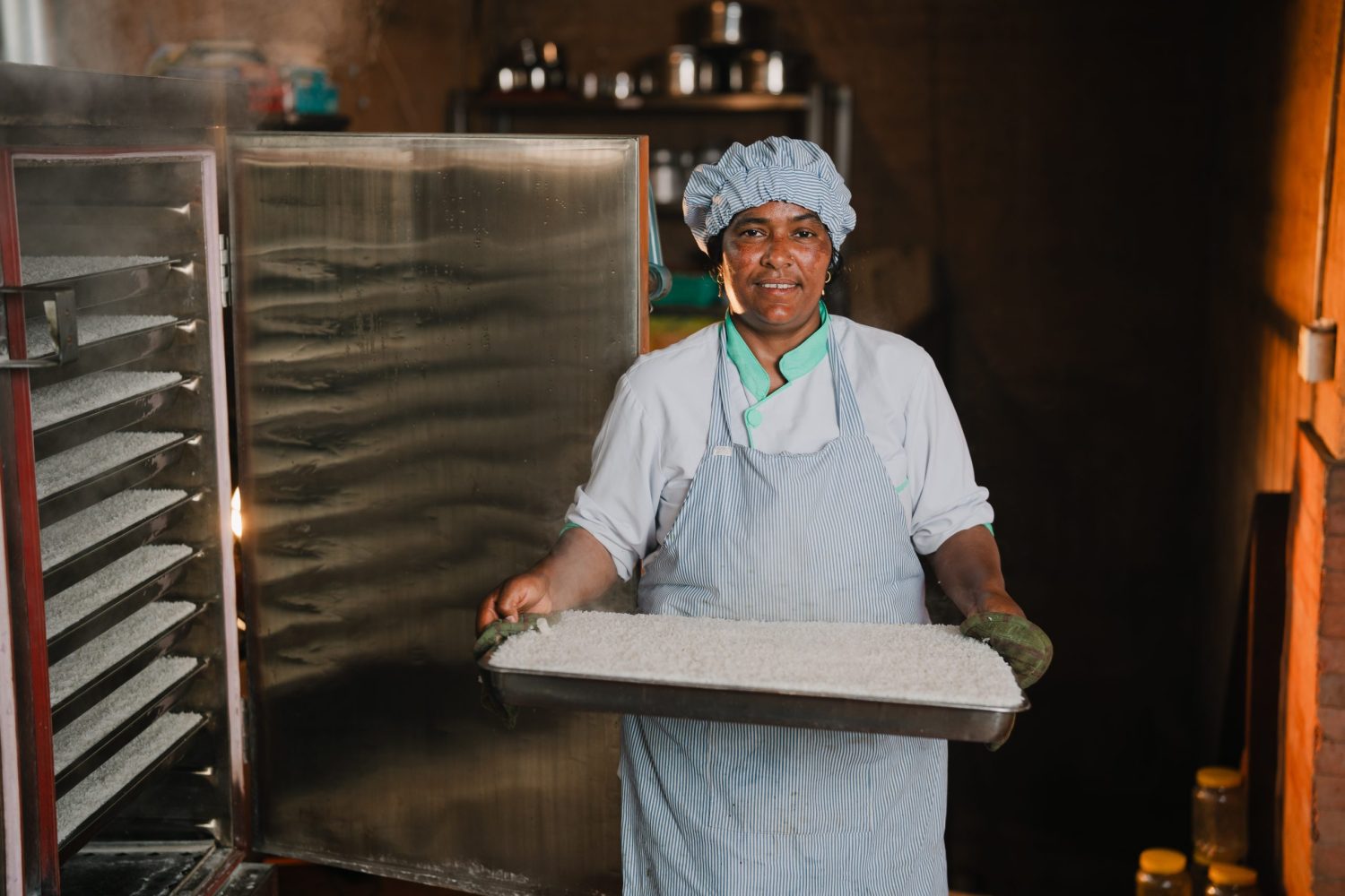 A woman wearing a food service head covering stands in a large commercial kitchen, holding a large pan of rice.
