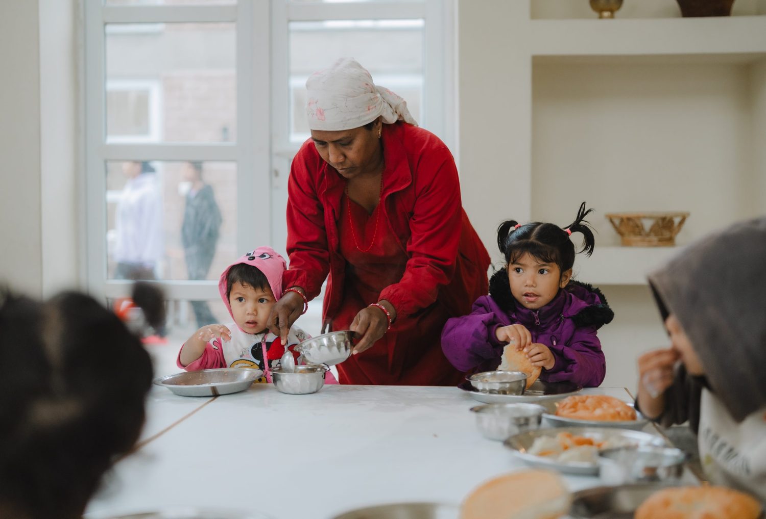A woman wearing a head scarf serves a meal to two small children at a table.