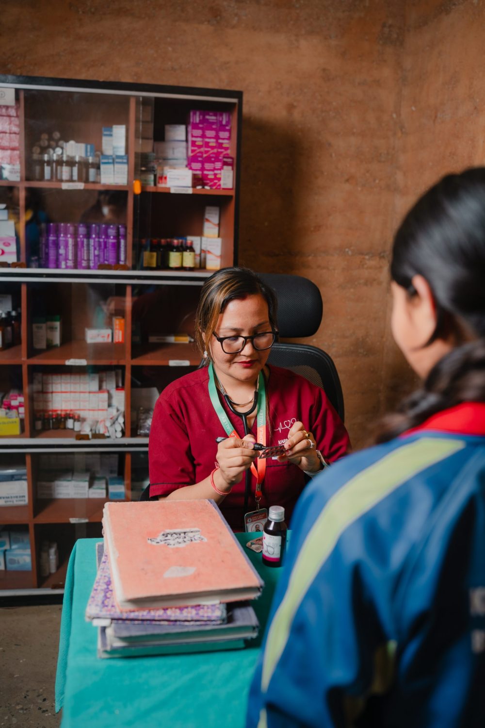 A nurse sits at a desk in a medical office with shelving with prescriptions on the wall. An adolescent girl stands in front of the desk facing away from the camera.