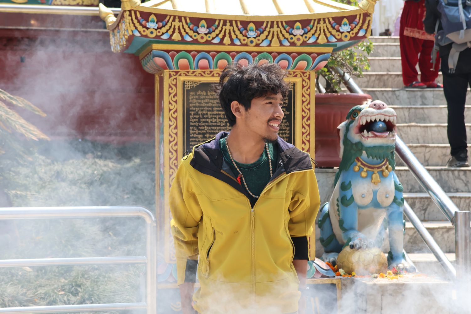 A young man in a yellow jacket stands in front of a gold-colored shrine next to a small statue of a dragon.