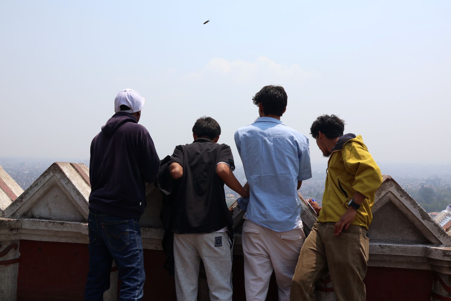 Four young men stand at the edge of a balcony and gaze out into a cloudy sky.