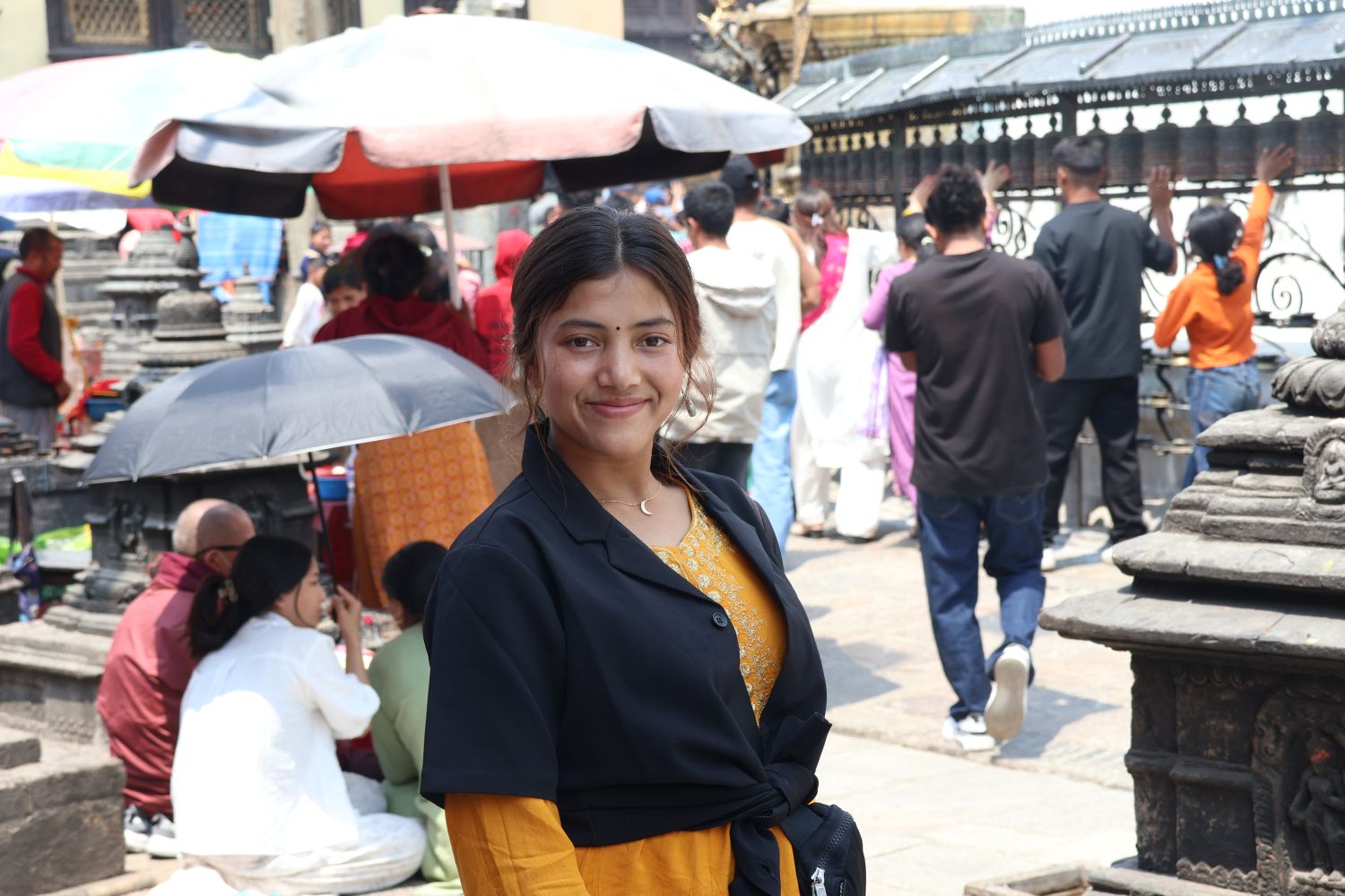 A young woman wearing a yellow dress and black cardigan sweater smiles in front of a street market.