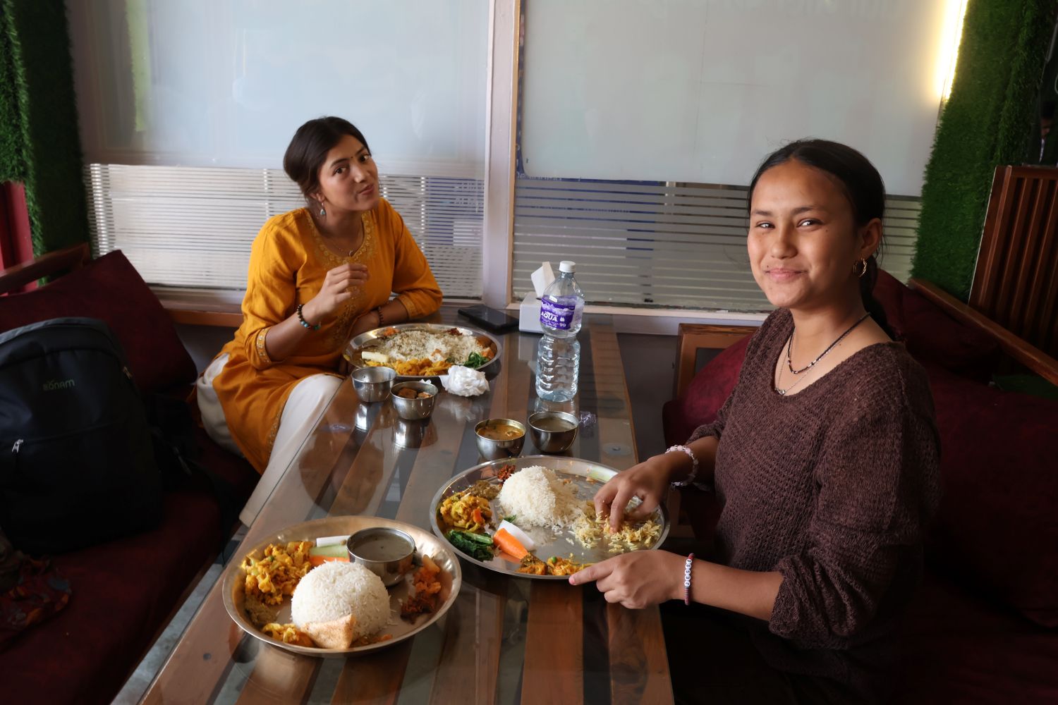Two young women sit at a table with traditional Nepali food on plates in front of them.