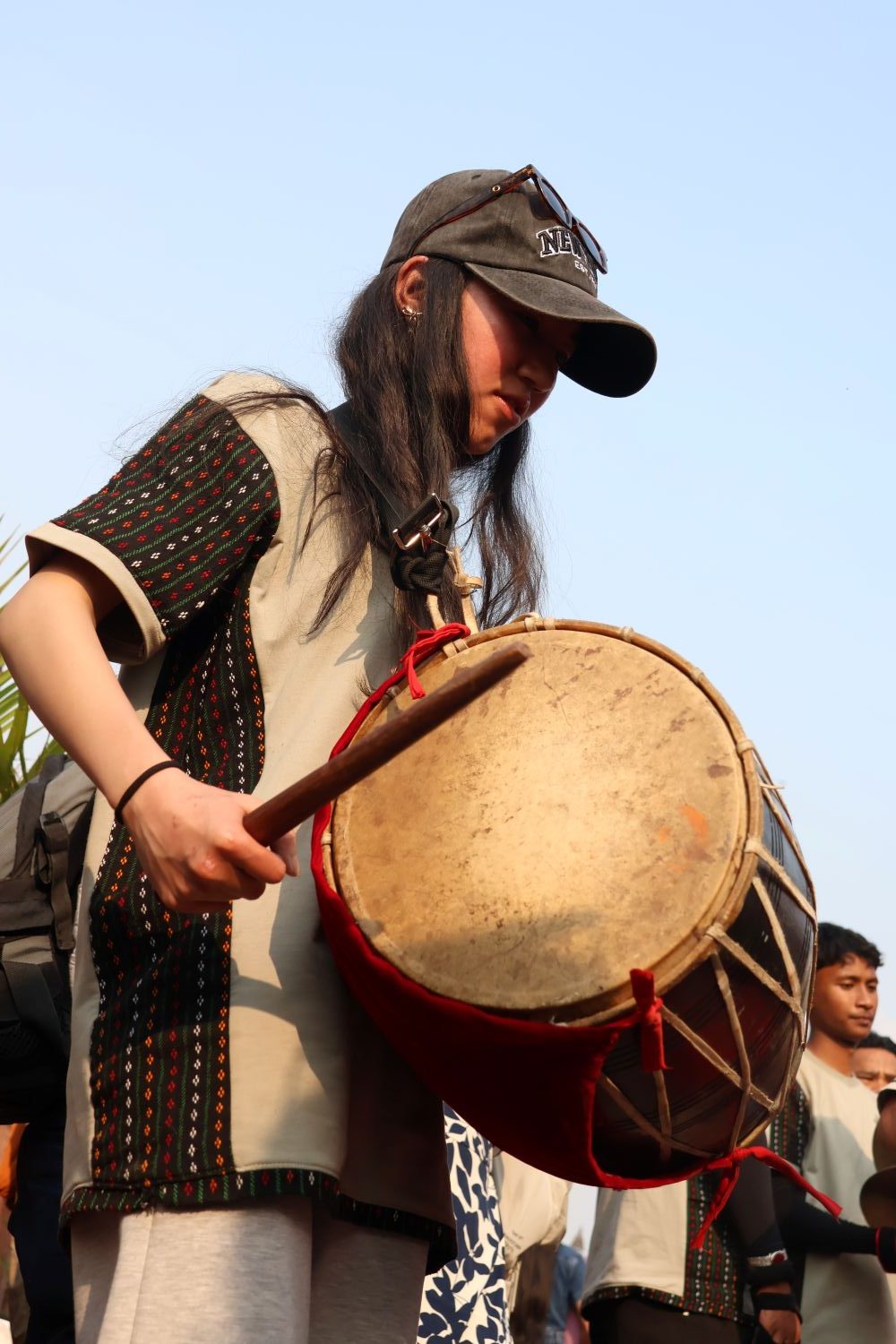 A person with long hair and a baseball cap beats a drum worn in front of them.
