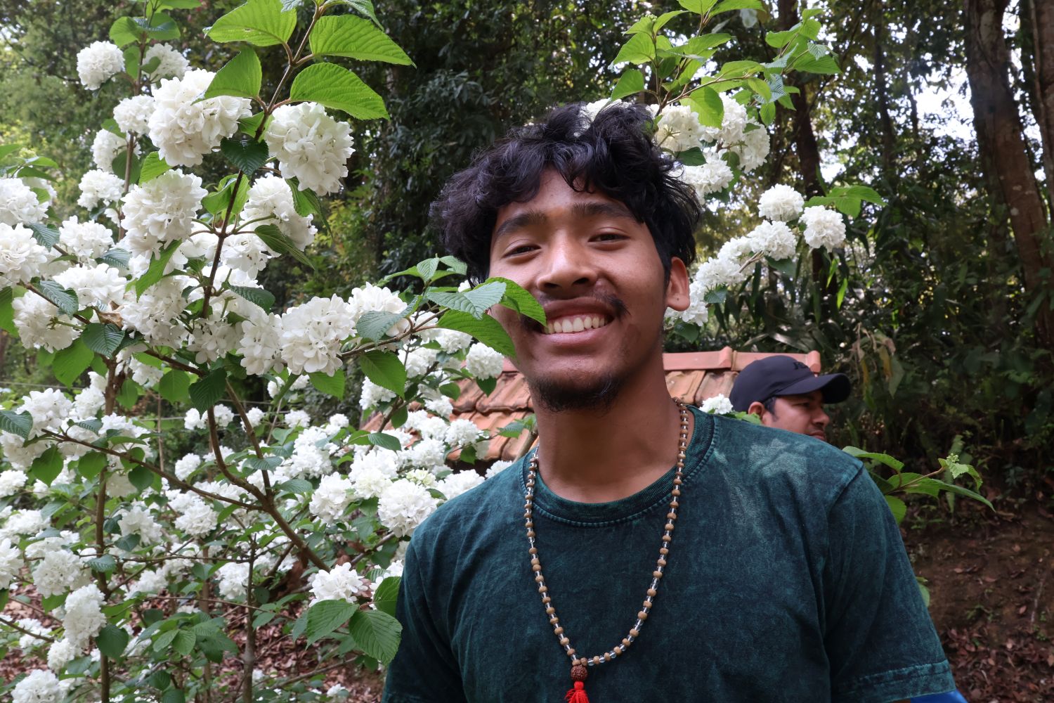 A young man stands next to a green tree with small white flowers.