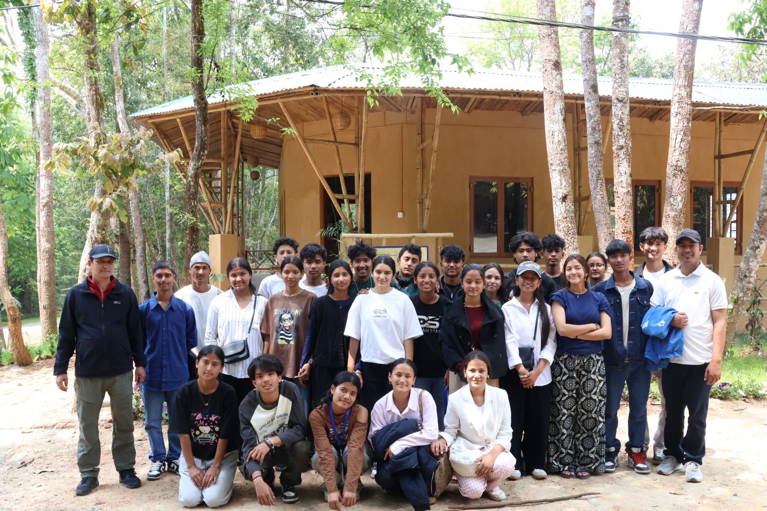 A group of students and adults pose in front of a building surrounded by green trees.