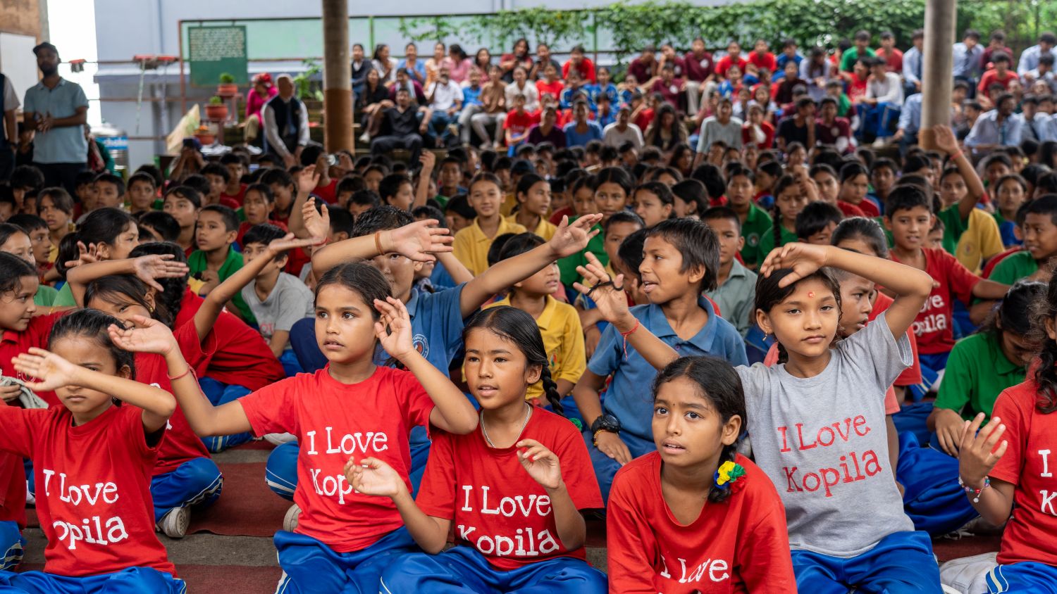 A group of students in red, blue, green, and yellow colorful "I Love Kopila" shirts raise their hands in the air, as if swaying to music.