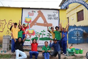 A group of students in colorful shirts pose in front of a mural with images of hands holding in collaboration. Images and a phrase in Nepal are painted on the mural and advocate for democracy, inclusion, and equality.
