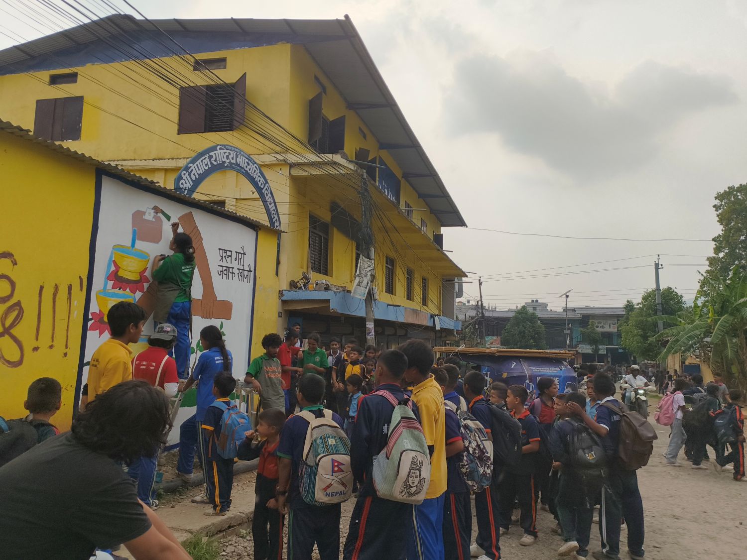 A group of students in colorful shirts watch as one student paints on a mural with images of hands holding in collaboration. Images and a phrase in Nepal are painted on the mural and advocate for democracy, inclusion, and equality.