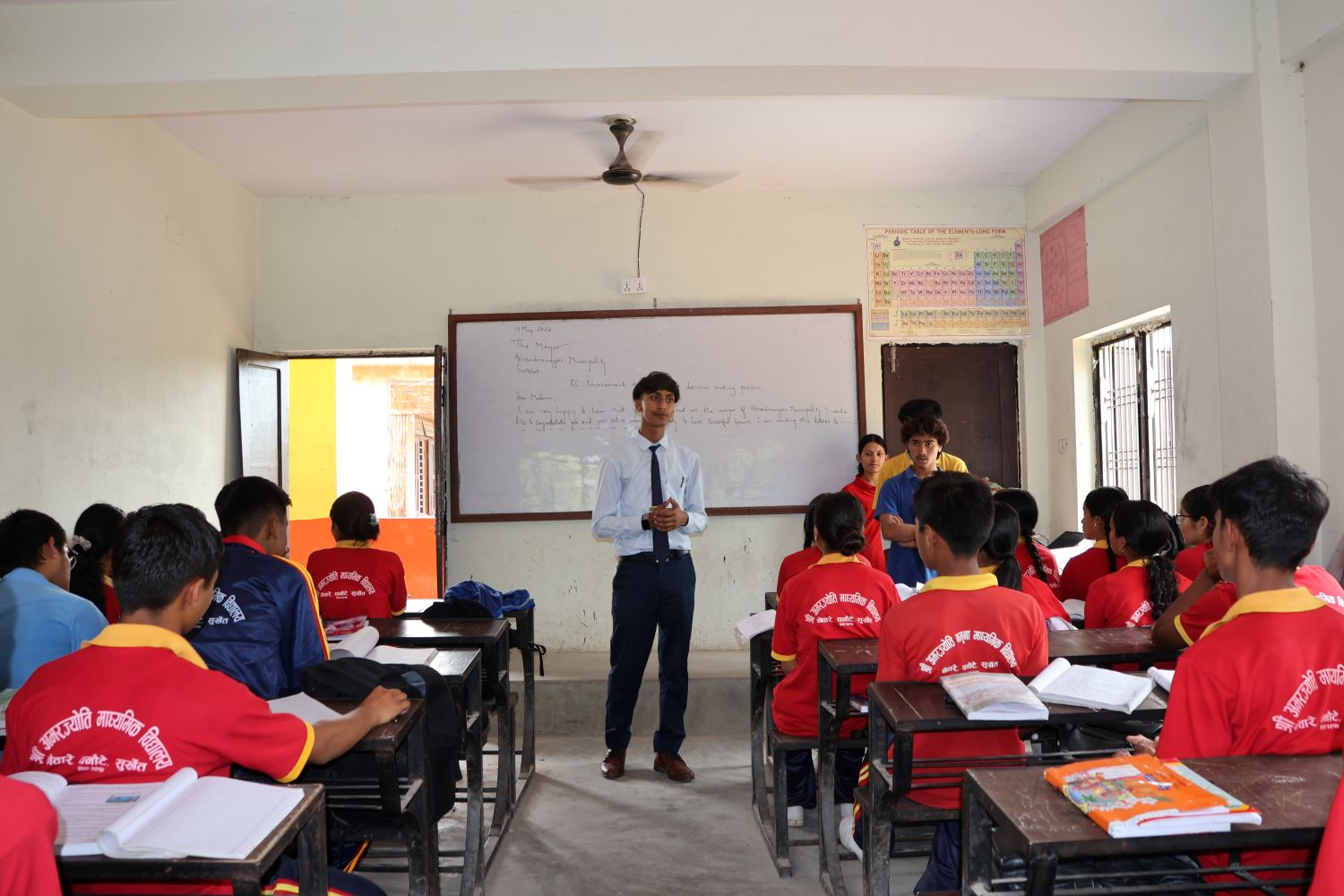 A student in a white dress shirt and tie stands at the front of a classroom. The students sitting at desks wear red school uniforms.