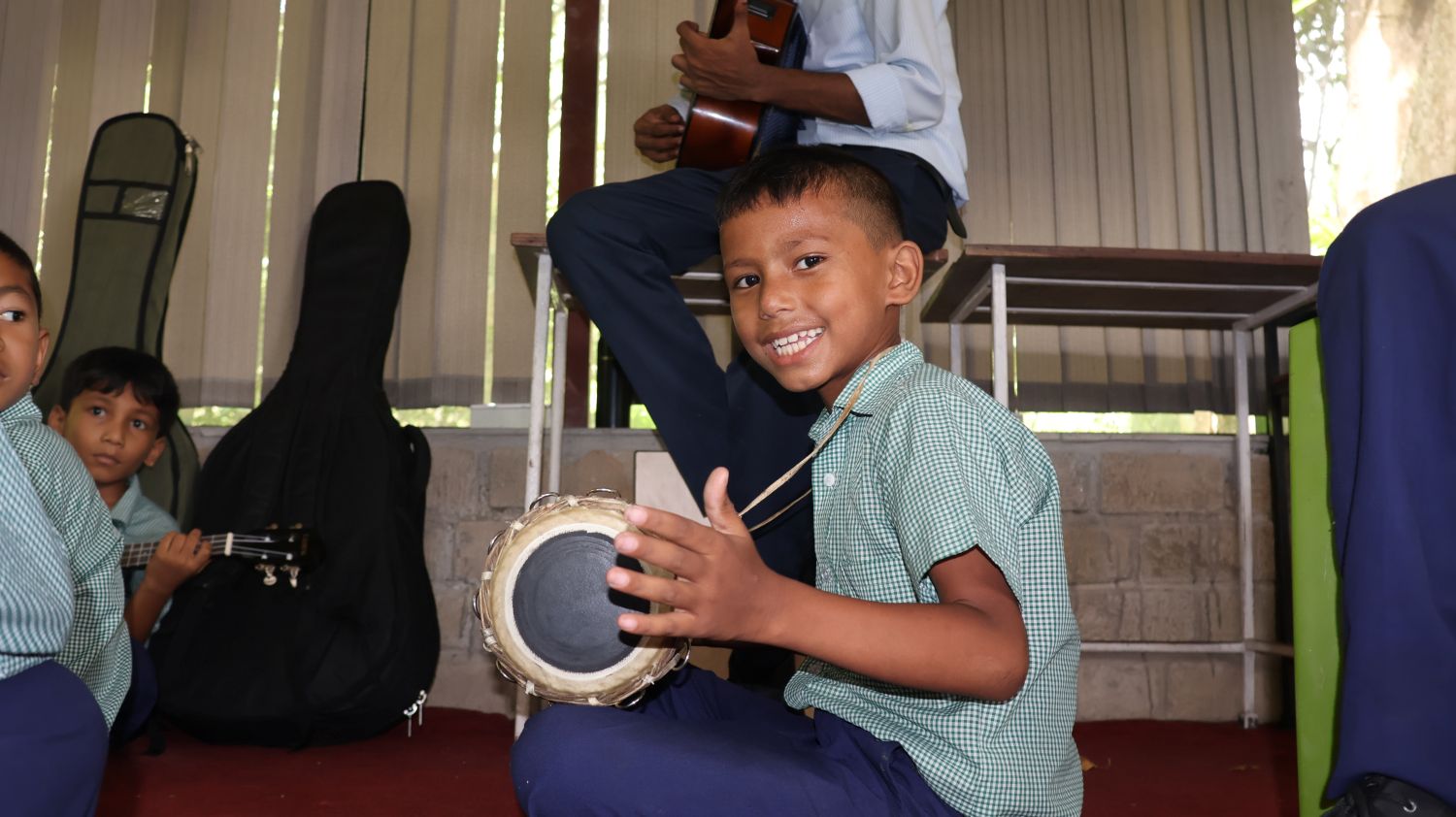 A boy in a school uniform holds a drum on his lap and smiles at the camera.