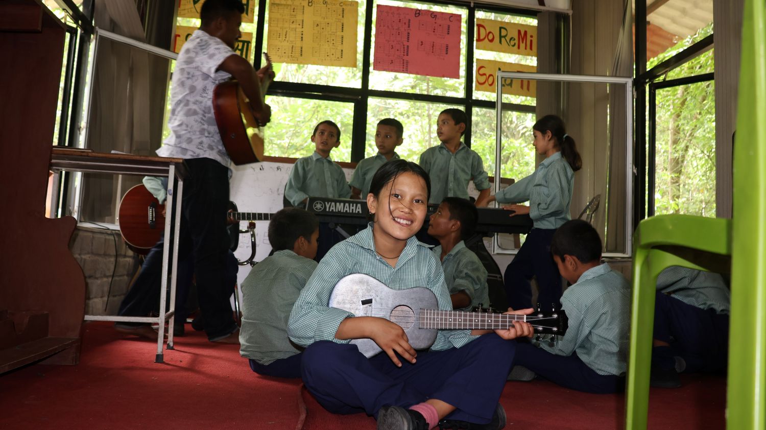 A young girl in a school uniform holds a small guitar and smiles at the camera. In the background, students and a teacher with instruments are practicing a song.