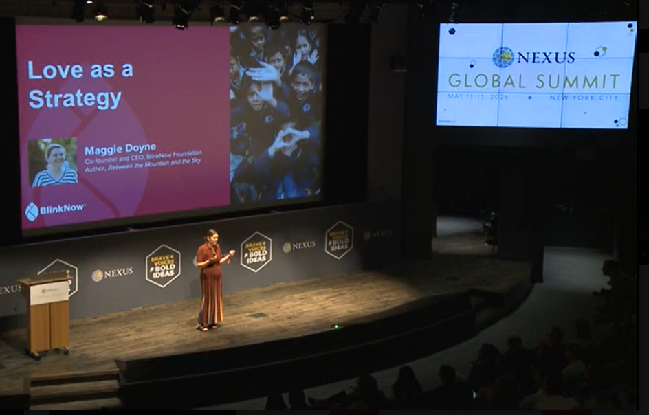 A woman stands on a stage making a presentation in front of a large screen with the title "Love as a Strategy."
