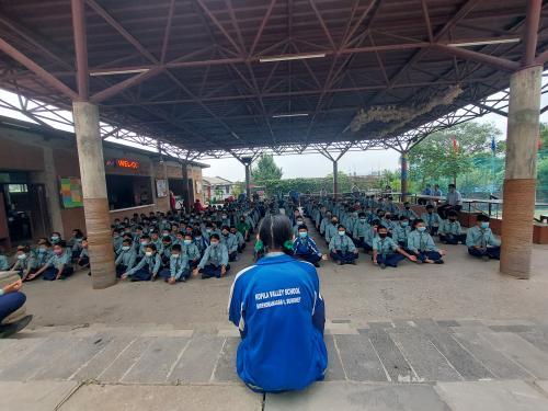 A student in a blue school shirt faces an audience of a hundred or more students in blue school uniforms. The students sit on the ground under an outdoor canopy.