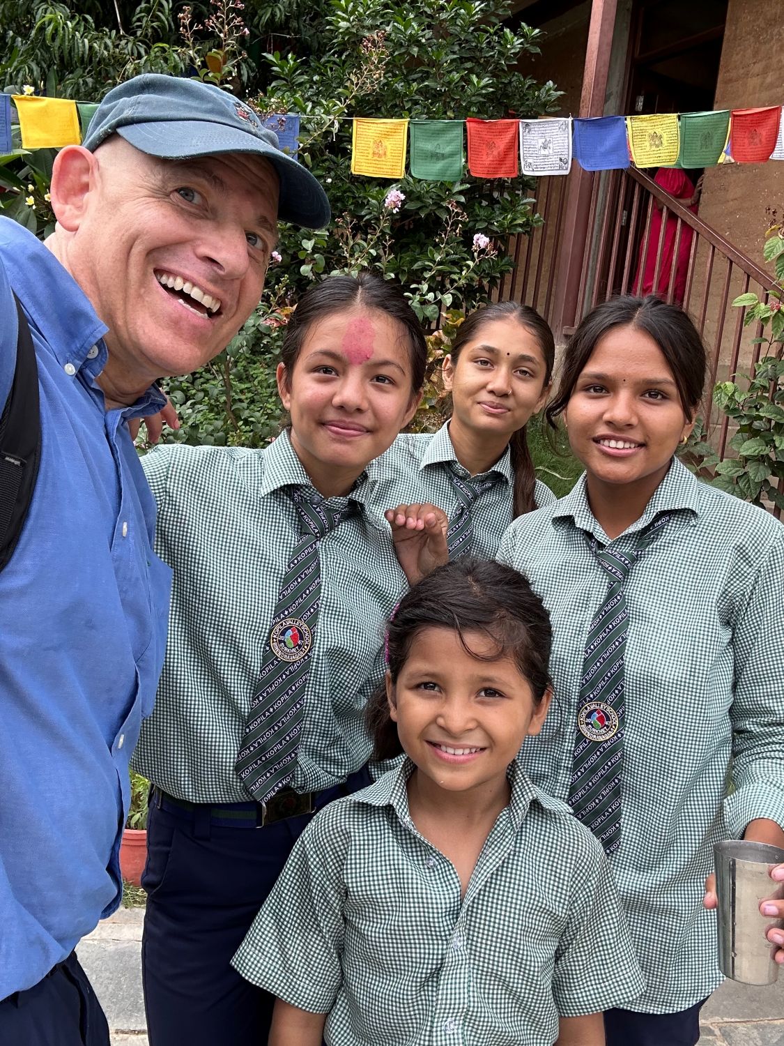 A man in a dress shirt and ball cap poses with some students in school uniforms. Colorful Nepali prayer flags hang in the background.