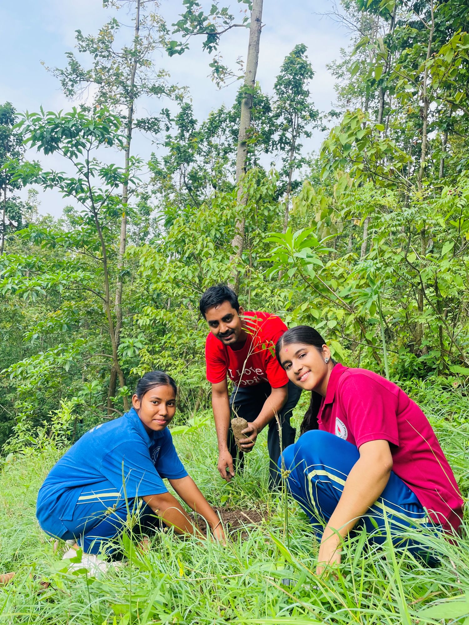 A teacher and two students in school polo shirts squat in some grass to plant small trees. The backdrop is a lush, green forest.