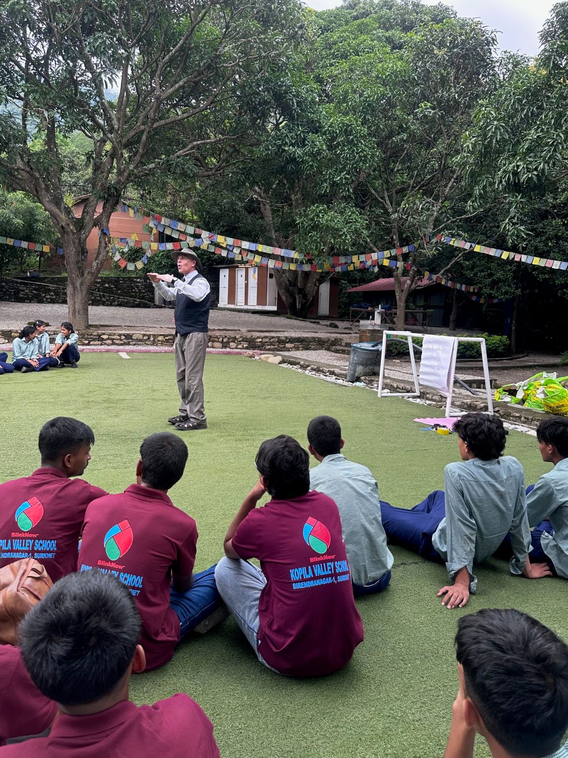 A man stands in front of a group of students in school uniforms sitting on an outdoor lawn. The background is lush green trees.