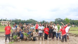 A group of students and teachers stand in a muddy rice field, covered in mud, with mountains in the background. Several people wear t-shirts that read "I love Kopila."