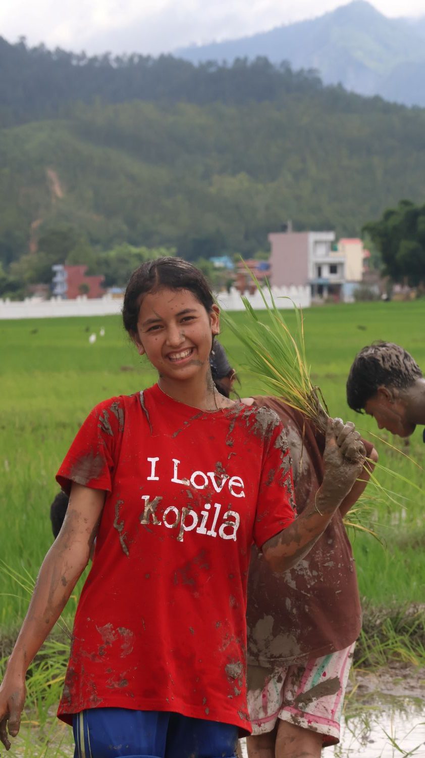 A teenage girl wearing a red "I Love Kopila" t-shirt holds a rice plant in front of a green field. Mountains are in the background.