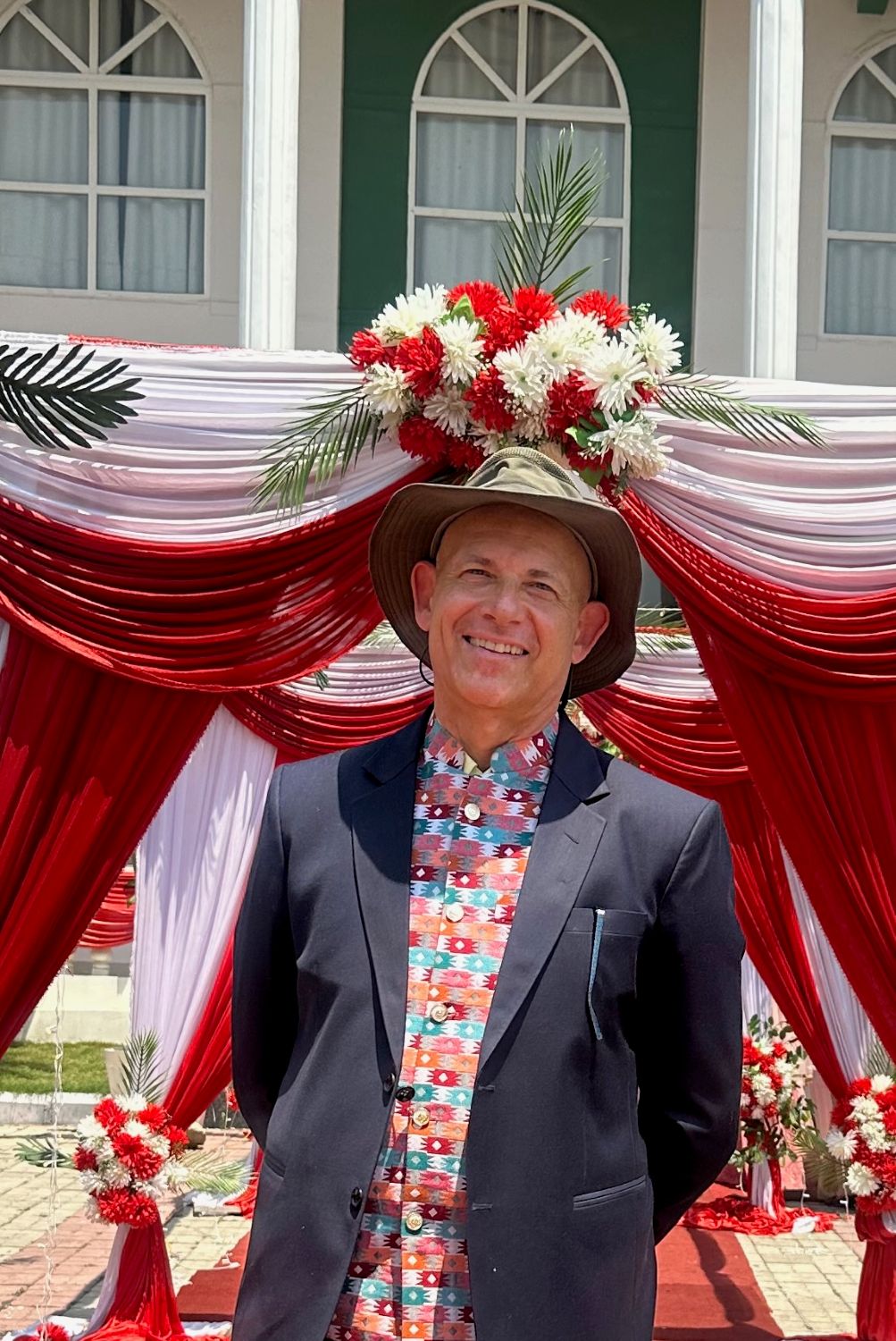 A man in a suit jacket and a hat stands in front of a white and red floral and drapery background.