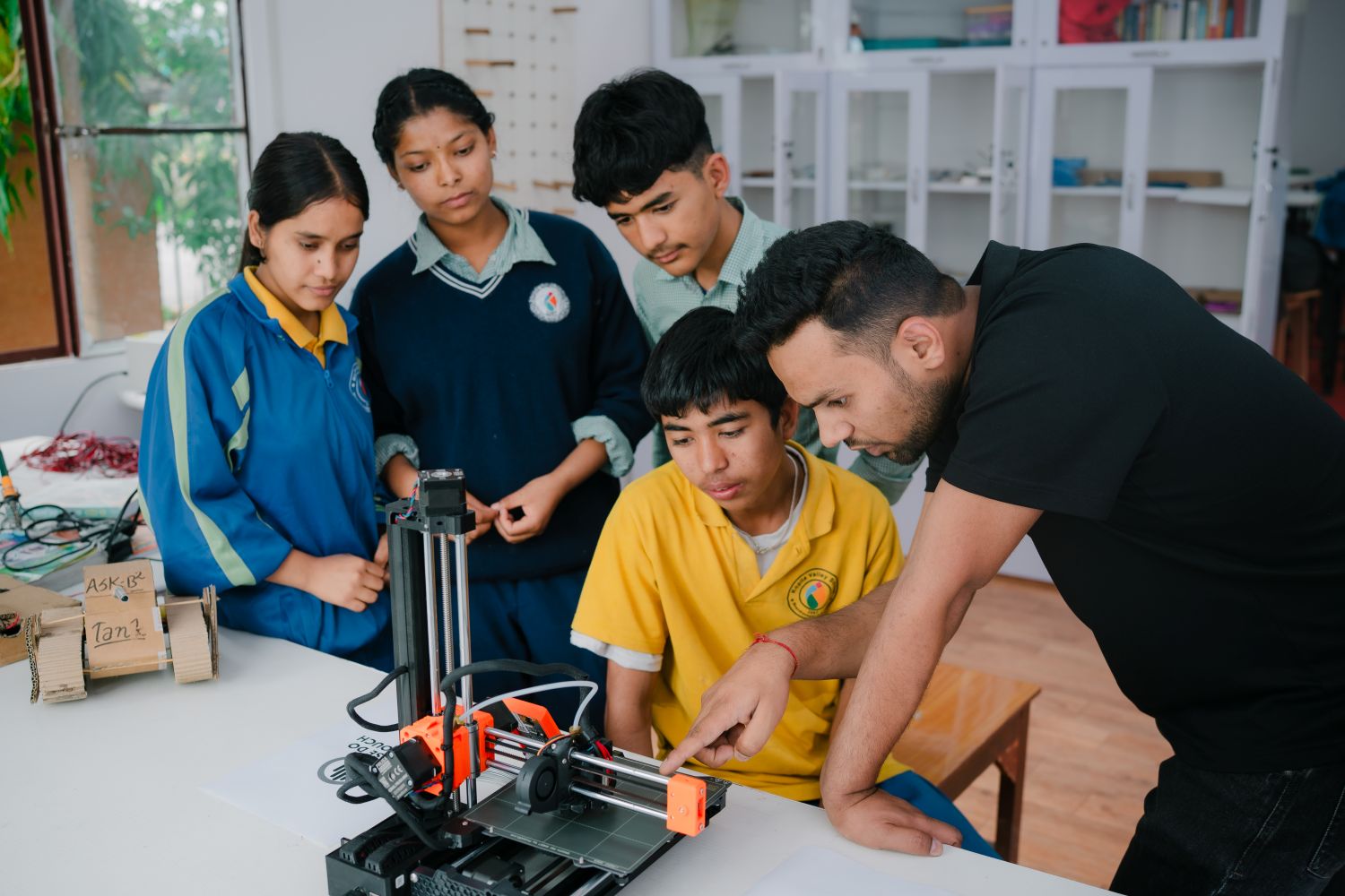 A group of students and a teacher lean over a white table with robotic equipment on it.